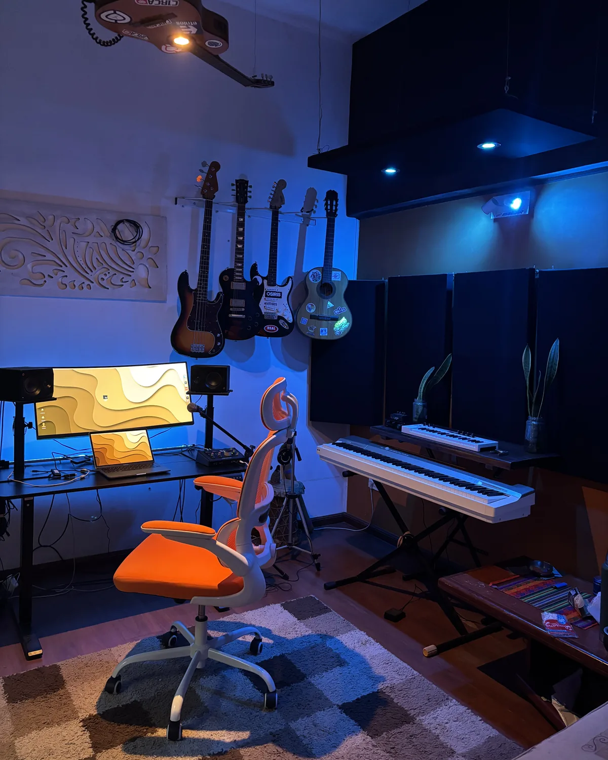 El estudio de ZECHE GRUV: escritorio con monitores, silla naranja, guitarras y bajo colgados en la pared, teclados, plantas y paneles acústicos bajo luz azul
